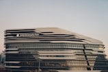 A modern architectural structure with a futuristic design, featuring sleek, curved lines and extensive glass panels. The building is identified as the Jockey Club Innovation Tower.