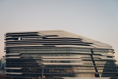 A modern architectural structure with a futuristic design, featuring sleek, curved lines and extensive glass panels. The building is identified as the Jockey Club Innovation Tower.