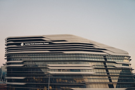 A modern architectural structure with a futuristic design, featuring sleek, curved lines and extensive glass panels. The building is identified as the Jockey Club Innovation Tower.