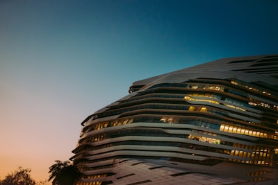 A modern, futuristic building with a unique, wavy architectural design is featured. The structure is illuminated, showcasing its horizontal layers and large glass windows. The sky transitions from a soft, warm sunset hue to a deeper blue, enhancing the building's elegant contours. Sparse silhouettes of trees are visible at the base, adding a natural element.