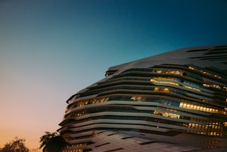 A modern, futuristic building with a unique, wavy architectural design is featured. The structure is illuminated, showcasing its horizontal layers and large glass windows. The sky transitions from a soft, warm sunset hue to a deeper blue, enhancing the building's elegant contours. Sparse silhouettes of trees are visible at the base, adding a natural element.