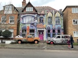 A vibrantly painted house featuring colorful murals and intricate designs covers the facade. The artwork includes abstract patterns, faces, and text reading 'I'm an artist your rules don't apply.' In front of the house are two vehicles, each adorned with unique artwork: a truck with orange and green patterns and a retro car with black and white designs. Two people stand on the sidewalk nearby, adding a touch of life to the scene.