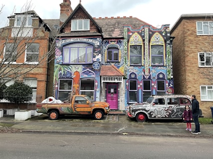 A vibrantly painted house featuring colorful murals and intricate designs covers the facade. The artwork includes abstract patterns, faces, and text reading 'I'm an artist your rules don't apply.' In front of the house are two vehicles, each adorned with unique artwork: a truck with orange and green patterns and a retro car with black and white designs. Two people stand on the sidewalk nearby, adding a touch of life to the scene.