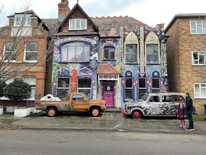A vibrantly painted house featuring colorful murals and intricate designs covers the facade. The artwork includes abstract patterns, faces, and text reading 'I'm an artist your rules don't apply.' In front of the house are two vehicles, each adorned with unique artwork: a truck with orange and green patterns and a retro car with black and white designs. Two people stand on the sidewalk nearby, adding a touch of life to the scene.