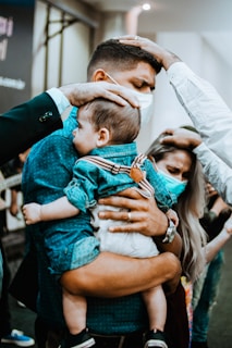 A man holding a baby is surrounded by people placing their hands on him, indicating a moment of prayer or support. Both the man and baby appear to be wearing masks. The presence of other hands suggests a communal or religious gathering.