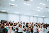 A large group of people gathered in a spacious, well-lit room. Many individuals have their arms raised, some appear to be singing or speaking. The crowd contains a diverse group of adults, with a number wearing face masks. The environment suggests a setting of engagement or worship.