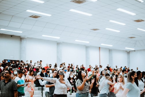 A large group of people gathered in a spacious, well-lit room. Many individuals have their arms raised, some appear to be singing or speaking. The crowd contains a diverse group of adults, with a number wearing face masks. The environment suggests a setting of engagement or worship.