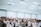 A large group of people gathered in a spacious, well-lit room. Many individuals have their arms raised, some appear to be singing or speaking. The crowd contains a diverse group of adults, with a number wearing face masks. The environment suggests a setting of engagement or worship.