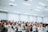 A large group of people gathered in a spacious, well-lit room. Many individuals have their arms raised, some appear to be singing or speaking. The crowd contains a diverse group of adults, with a number wearing face masks. The environment suggests a setting of engagement or worship.