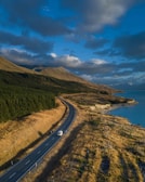 A winding road stretches through a scenic landscape featuring rolling hills and a forest on one side. A white van is traveling along the road, while a body of water lies on the opposite side. The sky above is filled with clouds and the light suggests a time close to sunset or sunrise.