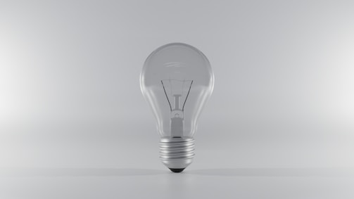 A clear, transparent light bulb standing upright on a minimalistic, light gray background. The bulb's glass and metal base are clearly visible, with the filament inside the bulb subtly highlighted.
