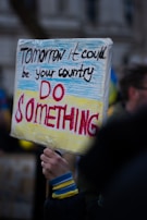 A protest sign with the words 'Tomorrow it could be your country, DO SOMETHING' written on it. The sign is held by a person wearing a black jacket with blue and yellow cuffs. The background is slightly blurred with other people and several flags.