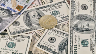 A collection of U.S. hundred dollar bills is scattered with a gold-colored Bitcoin coin placed on top, showcasing a contrast between traditional currency and cryptocurrency.