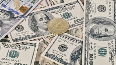 A collection of U.S. hundred dollar bills is scattered with a gold-colored Bitcoin coin placed on top, showcasing a contrast between traditional currency and cryptocurrency.