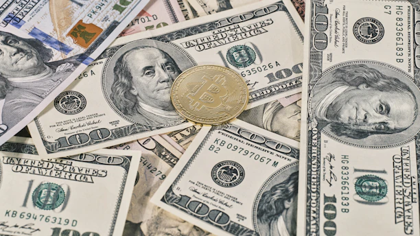 A collection of U.S. hundred dollar bills is scattered with a gold-colored Bitcoin coin placed on top, showcasing a contrast between traditional currency and cryptocurrency.
