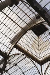 A glass and metal ceiling composed of multiple arch structures with intricate geometric patterns. The architectural design allows natural light to filter through, creating a bright and airy atmosphere. The metal framework holds large glass panels in place, and the overall structure appears to be part of an old, grand building.