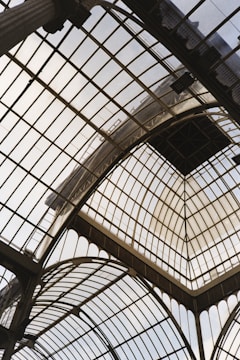 A glass and metal ceiling composed of multiple arch structures with intricate geometric patterns. The architectural design allows natural light to filter through, creating a bright and airy atmosphere. The metal framework holds large glass panels in place, and the overall structure appears to be part of an old, grand building.