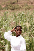 A person with braided hair adorned with beads stands confidently in a lush green cornfield, wearing a white outfit. The background features dense vegetation and a slightly blurred hilly terrain.