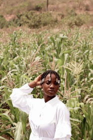 A person with braided hair adorned with beads stands confidently in a lush green cornfield, wearing a white outfit. The background features dense vegetation and a slightly blurred hilly terrain.