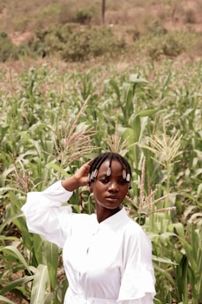 A person with braided hair adorned with beads stands confidently in a lush green cornfield, wearing a white outfit. The background features dense vegetation and a slightly blurred hilly terrain.