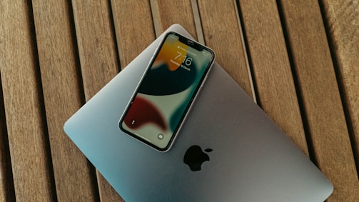 A smartphone rests on top of a closed laptop, both placed on a wooden surface. The phone's screen displays the time and date with a colorful background. The laptop's exterior features a prominent logo.