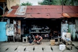 A person is working on a mechanical device outside a dilapidated workshop with a rusted corrugated metal roof. The structure is made of wooden planks with peeling paint and various tools and materials scattered around. A handwritten sign is affixed to a post, and there are several containers and buckets on a wooden table nearby.