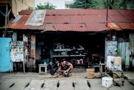A person is working on a mechanical device outside a dilapidated workshop with a rusted corrugated metal roof. The structure is made of wooden planks with peeling paint and various tools and materials scattered around. A handwritten sign is affixed to a post, and there are several containers and buckets on a wooden table nearby.