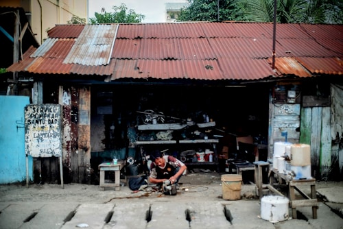 A person is working on a mechanical device outside a dilapidated workshop with a rusted corrugated metal roof. The structure is made of wooden planks with peeling paint and various tools and materials scattered around. A handwritten sign is affixed to a post, and there are several containers and buckets on a wooden table nearby.