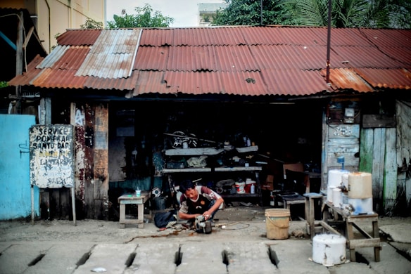 A person is working on a mechanical device outside a dilapidated workshop with a rusted corrugated metal roof. The structure is made of wooden planks with peeling paint and various tools and materials scattered around. A handwritten sign is affixed to a post, and there are several containers and buckets on a wooden table nearby.