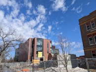 Modern building with a glass facade under construction or renovation, surrounded by fencing and construction debris. A tree with no leaves is visible in the foreground, along with a clear blue sky and scattered clouds.