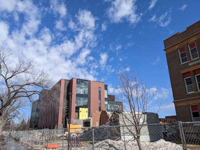 Modern building with a glass facade under construction or renovation, surrounded by fencing and construction debris. A tree with no leaves is visible in the foreground, along with a clear blue sky and scattered clouds.