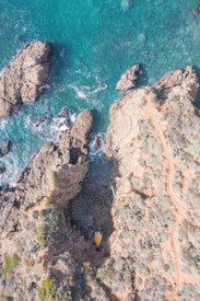 An aerial view of a rugged coastline with clear turquoise water. Large rocky formations jut into the sea, creating a natural cove. On the rocky terrain, several colorful surfboards are placed, adding a splash of color to the earthy landscape.