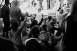 A group of people are gathered indoors in what appears to be a communal or religious setting. They are seated or kneeling, with many raising their hands in unison, indicating a moment of worship or collective experience. The scene is captured in black and white, adding a timeless quality to the image.