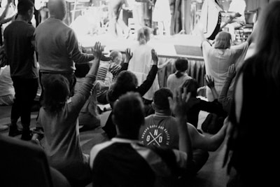 A group of people are gathered indoors in what appears to be a communal or religious setting. They are seated or kneeling, with many raising their hands in unison, indicating a moment of worship or collective experience. The scene is captured in black and white, adding a timeless quality to the image.