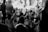 A group of people are gathered indoors in what appears to be a communal or religious setting. They are seated or kneeling, with many raising their hands in unison, indicating a moment of worship or collective experience. The scene is captured in black and white, adding a timeless quality to the image.