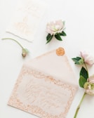 An elegant envelope with intricate calligraphy lies open, featuring ornate designs and a wax seal. Nearby are delicate flowers with soft pink petals and green leaves, complementing the overall romantic theme.
