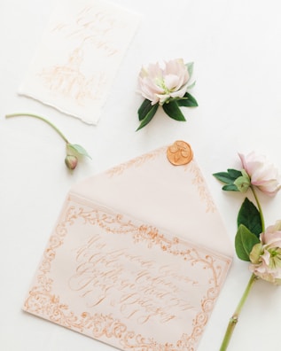 An elegant envelope with intricate calligraphy lies open, featuring ornate designs and a wax seal. Nearby are delicate flowers with soft pink petals and green leaves, complementing the overall romantic theme.
