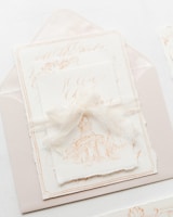 An elegant and artistic invitation card with a harmonious presentation. The card features a delicate illustration of a classical building, possibly a chapel or cathedral. It is placed on top of an envelope with fine calligraphy in soft, muted tones. A translucent ribbon is tied around the card, adding a touch of sophistication and charm.