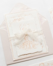 An elegant and artistic invitation card with a harmonious presentation. The card features a delicate illustration of a classical building, possibly a chapel or cathedral. It is placed on top of an envelope with fine calligraphy in soft, muted tones. A translucent ribbon is tied around the card, adding a touch of sophistication and charm.