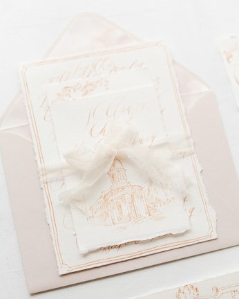 An elegant and artistic invitation card with a harmonious presentation. The card features a delicate illustration of a classical building, possibly a chapel or cathedral. It is placed on top of an envelope with fine calligraphy in soft, muted tones. A translucent ribbon is tied around the card, adding a touch of sophistication and charm.