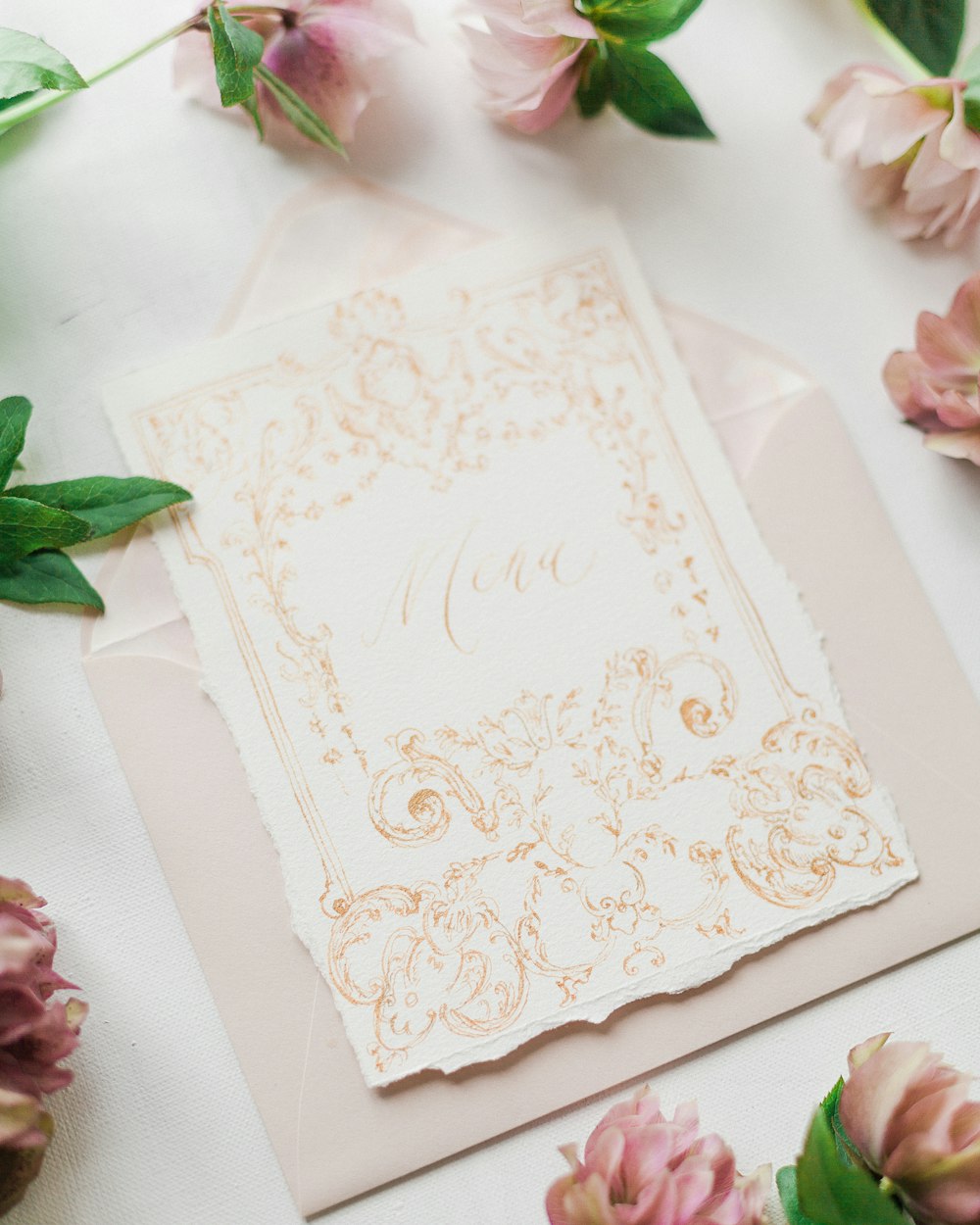 Una tarjeta de boda blanca y dorada en una mesa con flores rosadas