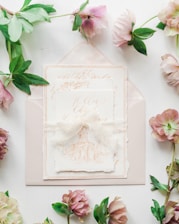 A set of elegant wedding invitations featuring intricate calligraphy and an illustration of a building sits atop an envelope. Surrounding the invitations, delicate flowers and green leaves add a touch of nature.