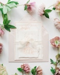 A set of elegant wedding invitations featuring intricate calligraphy and an illustration of a building sits atop an envelope. Surrounding the invitations, delicate flowers and green leaves add a touch of nature.