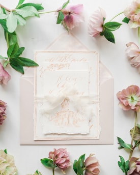 A set of elegant wedding invitations featuring intricate calligraphy and an illustration of a building sits atop an envelope. Surrounding the invitations, delicate flowers and green leaves add a touch of nature.