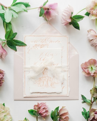 A set of elegant wedding invitations featuring intricate calligraphy and an illustration of a building sits atop an envelope. Surrounding the invitations, delicate flowers and green leaves add a touch of nature.