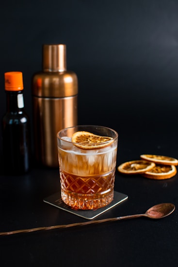 A crystal glass filled with an amber-colored cocktail garnished with a dried citrus slice sits on a dark coaster. Nearby, a copper cocktail shaker and a small bottle with an orange cap are visible. In the foreground, there is a copper spoon, and a few dried citrus slices are scattered next to the glass. The background is dark, creating a sophisticated and elegant atmosphere.