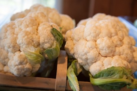 Several large cauliflower heads with light green leaves are placed in a wooden crate. The texture of the cauliflower is prominent, with compact, white florets.