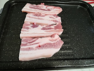 Three slices of raw pork belly are neatly arranged on a dark, speckled cooking tray. The meat has visible layers of pink flesh and white fat.