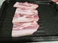 Three slices of raw pork belly are neatly arranged on a dark, speckled cooking tray. The meat has visible layers of pink flesh and white fat.