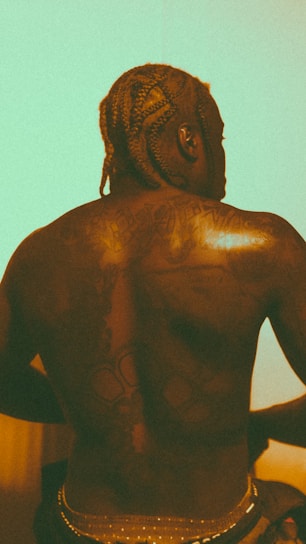 A person with braided hair seen from the back. The back is tattooed with various designs and the lighting gives a warm, vintage aesthetic.